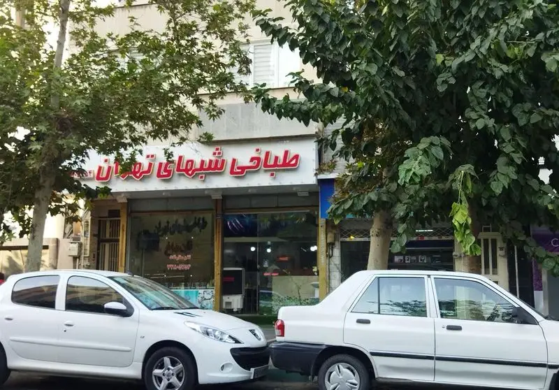تصویری از نمای بیرونی مغازه طباخی شبهای تهران با تابلو قرمز رنگ، در خیابانی پردرخت با دو خودروی سفید پارک شده در جلو