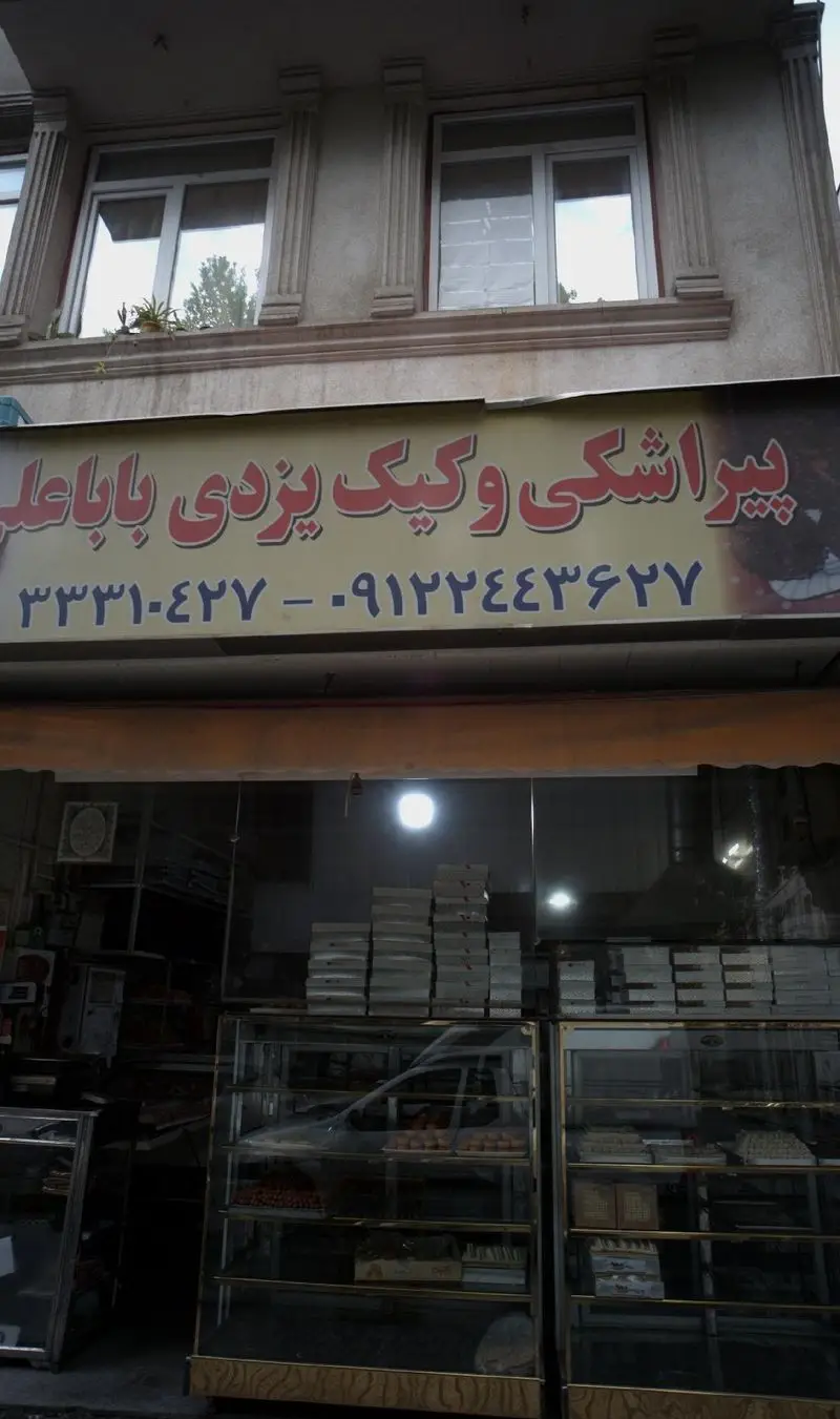 کسب و کار پیراشکی و کیک یزدی باباعلی با تابلوی بزرگ حاوی نام و شماره تماس، و نمای ویترین مملو از شیرینی‌های سنتی و مدرن.