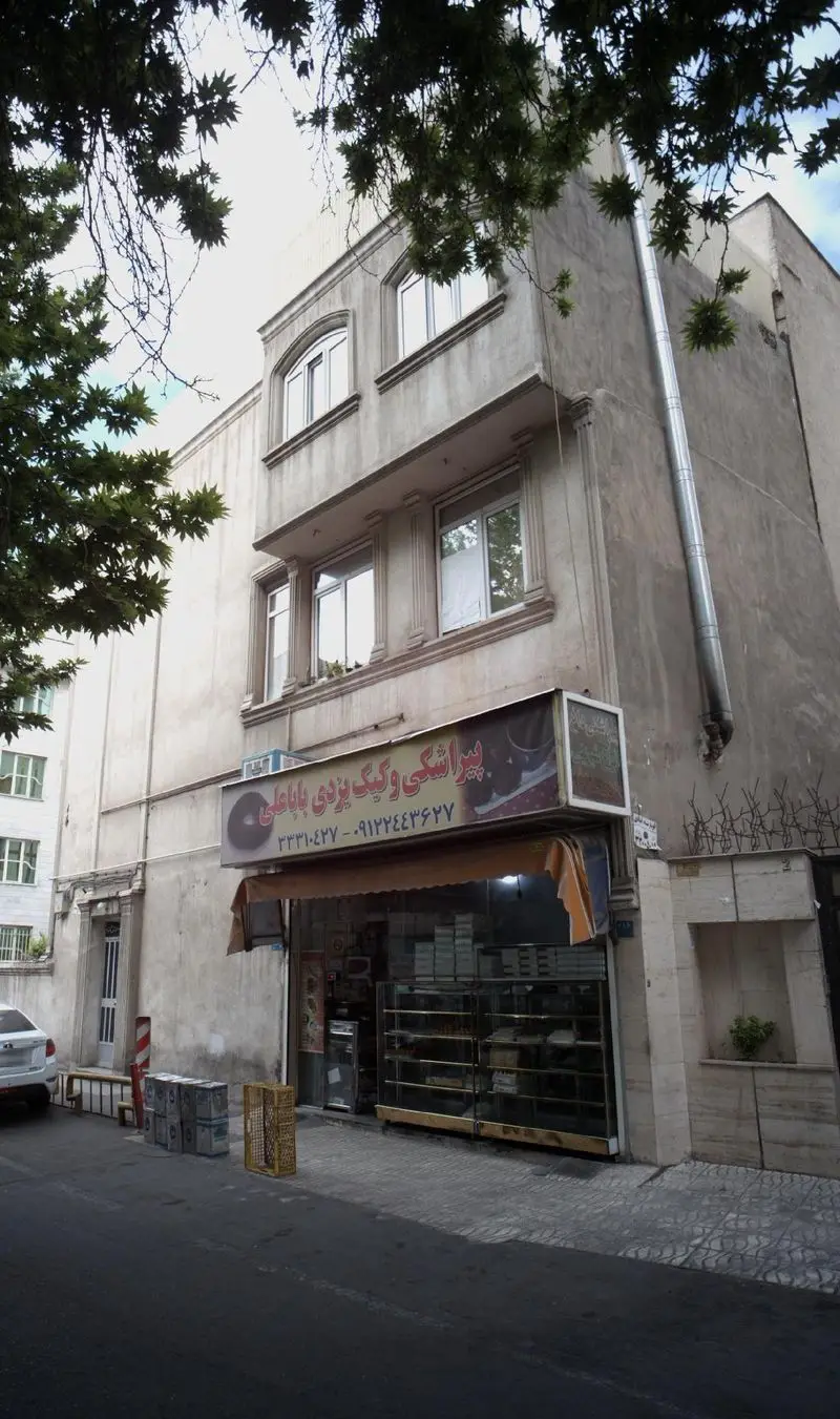 نمایی کامل از فروشگاه پیراشکی و کیک یزدی باباعلی، مرکزی برای تهیه شیرینی‌های سنتی و تازه، همراه با نمای شهری اطراف این کسب و کار.