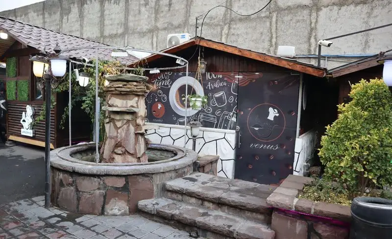 فضای باز دلنشین باغچه رستوران ونوس شهران با فواره سنگی، کافه و دکوراسیون قهوه. مناسب برای دورهمی و لذت بردن از قهوه و غذا.