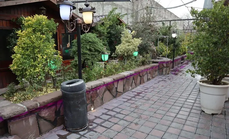 فضای دلنشین و سرسبز باغچه رستوران ونوس شهران با مسیر سنگفرش، دیوار سنگی، گیاهان متنوع و نورپردازی شبانه.