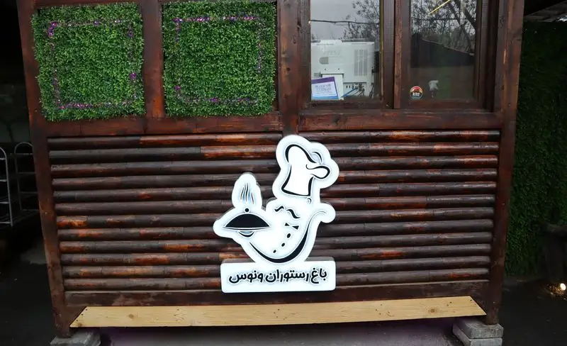 تابلوی ورودی باغچه رستوران ونوس شهران با لوگوی سفید سرآشپز و نام "باغ رستوران ونوس"، بر روی نمای چوبی و تزئینات سبز دیواری.