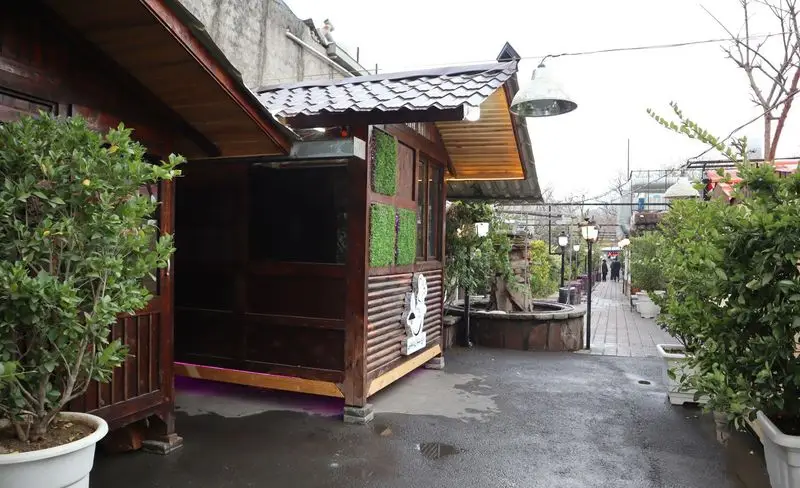 تصویری از فضای بیرونی باغچه رستوران ونوس شهران با آلاچیق‌های چوبی، گلدان‌های سبز و مسیر پیاده‌روی روشن، محیطی آرام و دلنشین.