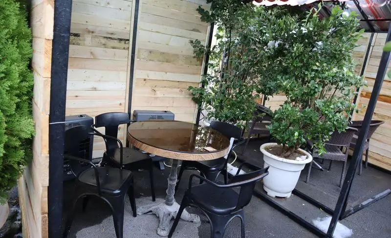 تصویر فضای باز باغچه رستوران ونوس شهران با میز گرد و صندلی‌های مشکی، بخاری‌ها و گلدان‌های سبز برای محیطی گرم و دلپذیر.