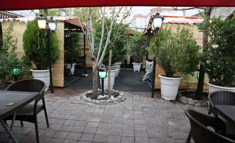 باغچه رستوران ونوس شهران: محیط سرسبز و آرام با آلاچیق‌های چوبی، میز و صندلی‌های حصیری، گلدان‌ها و درختان زیبا، فضایی دنج و دلنشین.