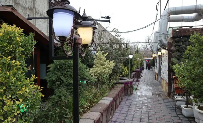 باغچه رستوران ونوس شهران با مسیر سنگفرش، فضای سبز دلنشین و چراغ‌های تزئینی، محیطی آرام و زیبا برای مشتریان در فضای باز.