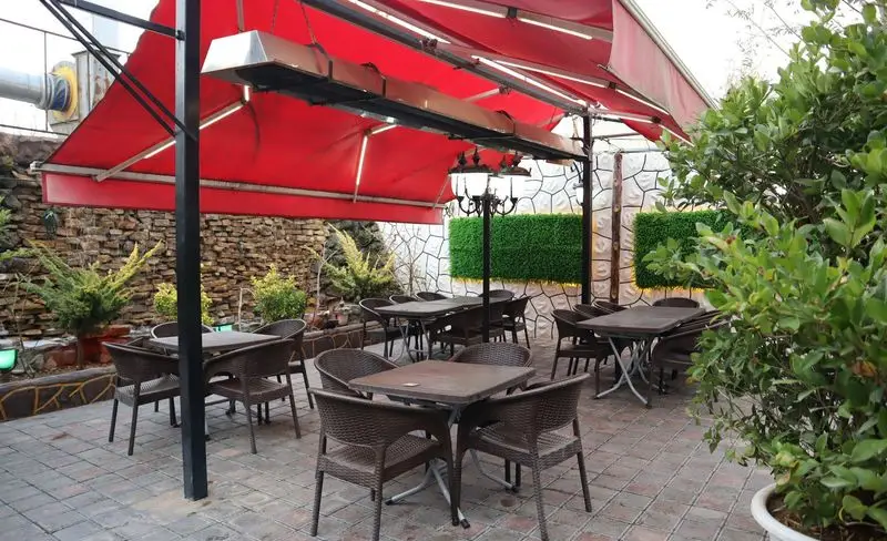 فضای بیرونی باغچه رستوران ونوس شهران با سایبان قرمز، هیتر، میز و صندلی‌های شیک، دیوار سنگی و فضای سبز دلپذیر.