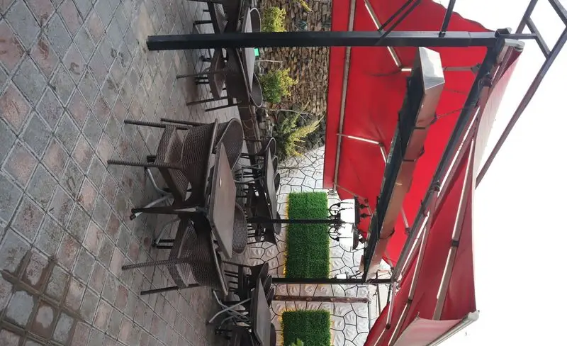 فضای باز باغچه رستوران ونوس شهران با میز و صندلی‌های شیک زیر سایبان قرمز، ایده‌آل برای صرف غذا و دورهمی در محیطی دلنشین.