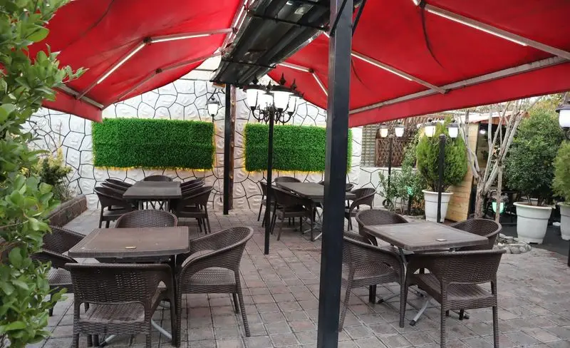 فضای باز دلنشین باغچه رستوران ونوس شهران تهران با سایبان‌های قرمز، میز و صندلی حصیری، دیوار سبز و نورپردازی زیبا.