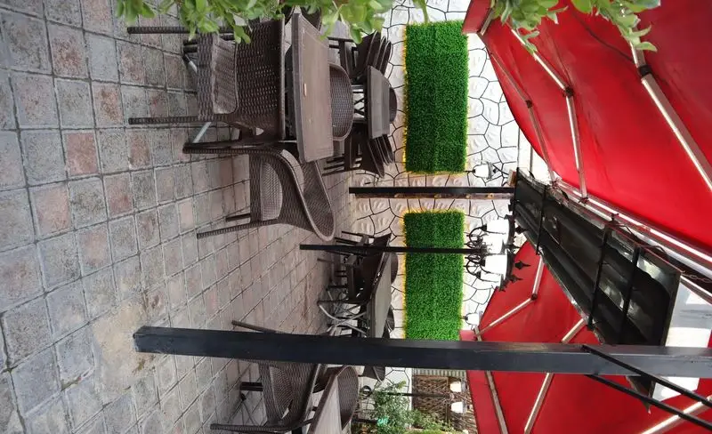 فضای باز دلنشین باغچه رستوران ونوس شهران با میز و صندلی حصیری، کفپوش سنگی، دیوارهای سبز و سایبان‌های قرمز