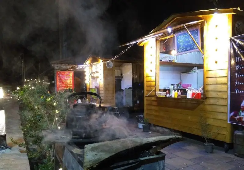 پیتزا رادیکال: منظره‌ای شبانه از دکه‌های غذای خیابانی در فضای باز با نورپردازی گرم، کتری بخار روی زغال و منو.