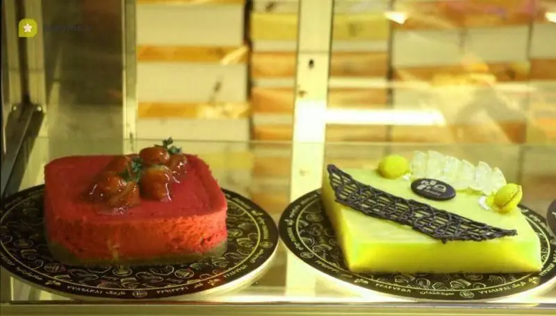 کیک‌های خوش‌طعم و جذاب نان و شیرینی تهران: چیزکیک توت فرنگی تازه و کیک سبز خاص با تزئینات زیبا در ویترین شیشه‌ای.