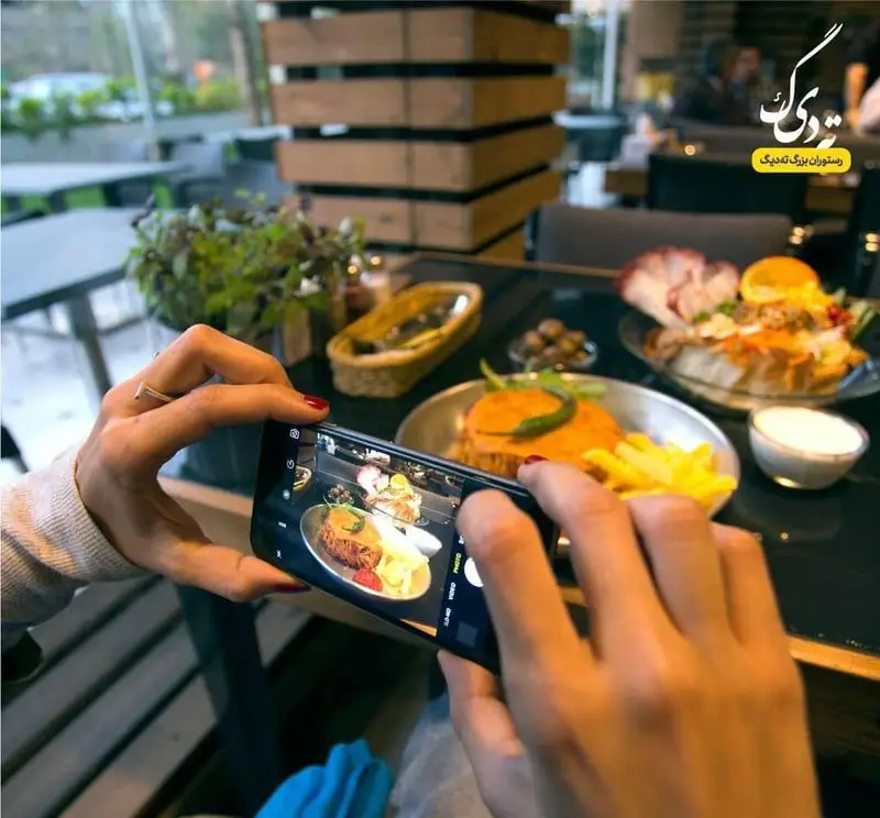 مشتری در رستوران ته دیگ در حال عکاسی از غذای لذیذ با موبایل. تجربه دلنشین و فضای مدرن رستوران ته دیگ برای مشتریان.