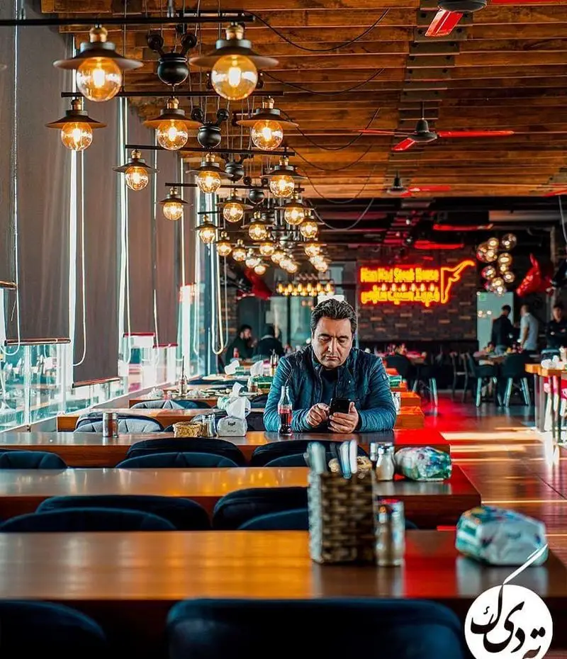 فضای داخلی رستوران ته دیگ با مشتری در حال مشاهده گوشی، سقف چوبی، لوسترهای زیبا و دکوراسیون مدرن و دلنشین