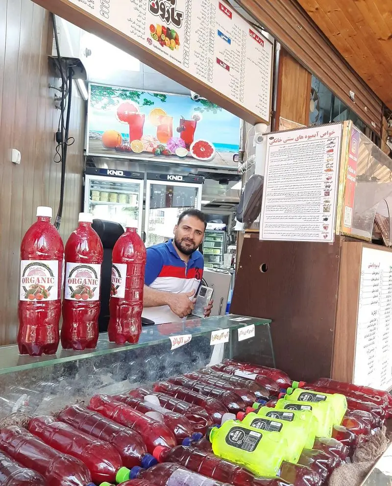 مغازه آبمیوه بستنی لب کارون با مردی فروشنده و ردیف‌های آبمیوه‌های طبیعی و ارگانیک، شامل شربت‌های آلبالو و میوه‌ای.