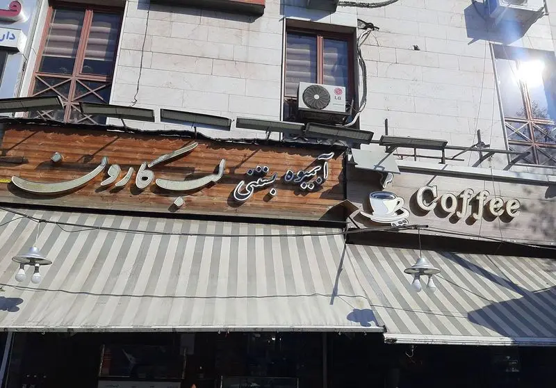 نمای خارجی از مغازه آبمیوه و بستنی لب کارون با تابلوی طلایی رنگ، همراه با تابلوی "Coffee" و سایبان راه راه سفید و خاکستری، زیر نور خورشید.