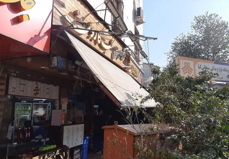 تصویر نمای خارجی مغازه آبمیوه و بستنی لب کارون، با تابلو و سایبان، به همراه قسمتی از منوی نوشیدنی‌ها و دسرهای خنک.