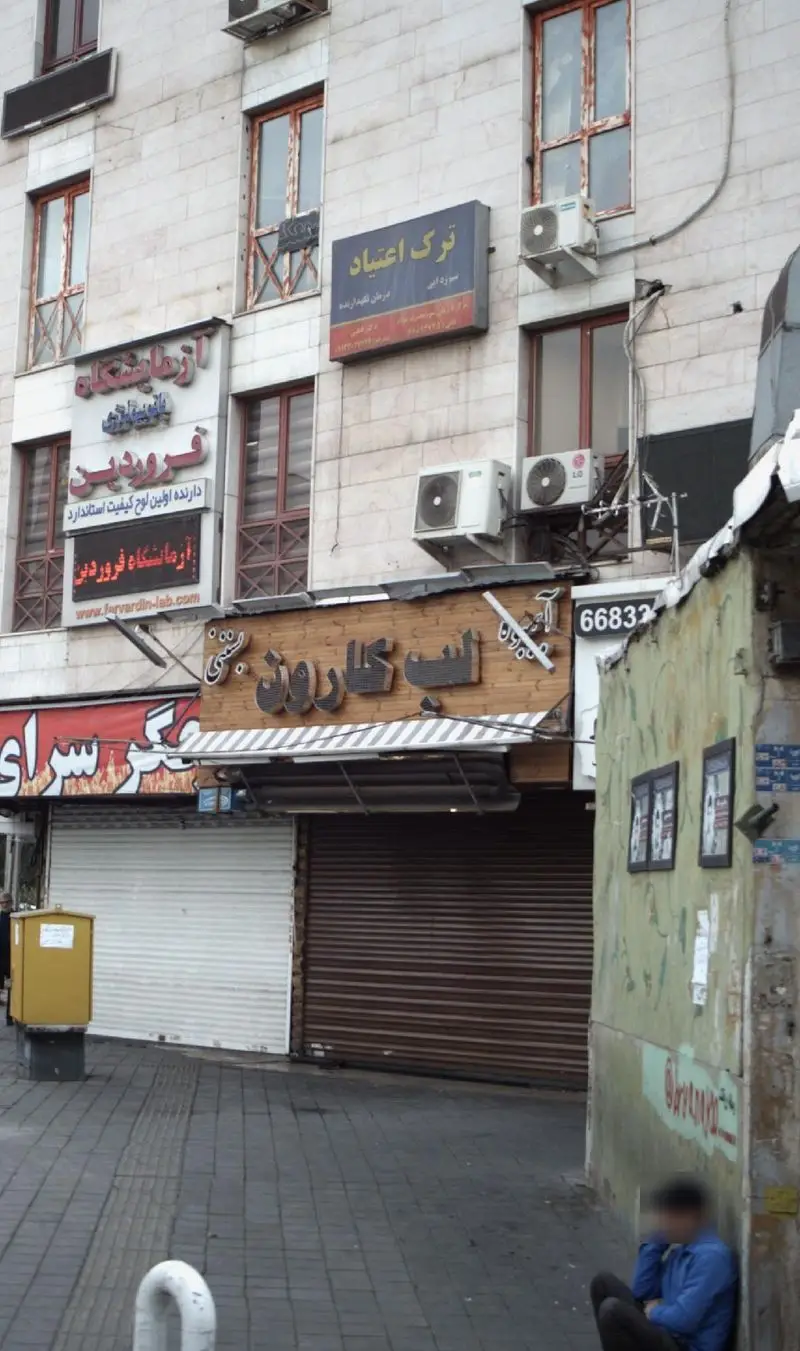 نمای بیرونی آبمیوه بستنی لب کارون در خیابان، با تابلوهای آزمایشگاه فروردین و ترک اعتیاد، و یک نفر نشسته روی پیاده‌رو.