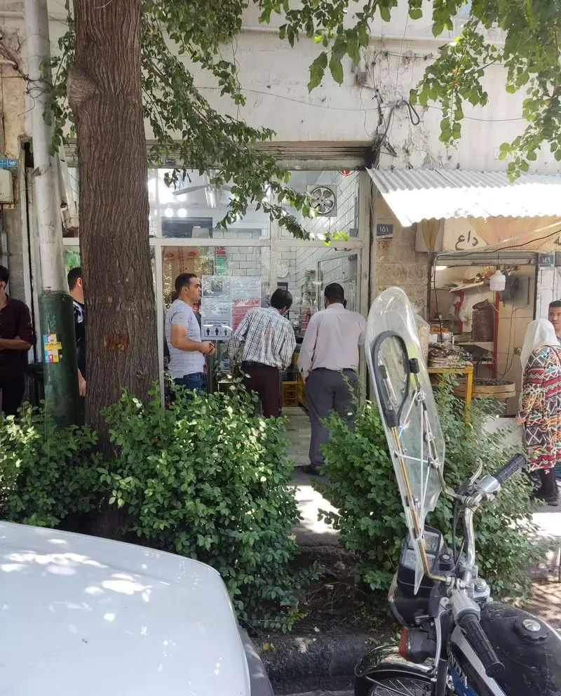 نمایی از ساندویچ موسیو در خیابان، با مشتریان و رهگذران. مغازه در محیطی شلوغ با درختان، موتور و خودرو در جلوی ورودی قرار دارد.