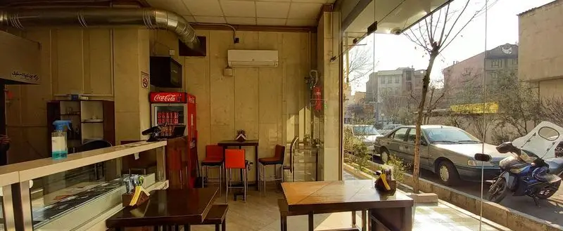 نمایی از داخل رستوران گردسوز با چیدمان ساده و گرم، میزهای چوبی، پیشخوان سفارشات و ویوی زیبای خیابان شهر از پنجره بزرگ.