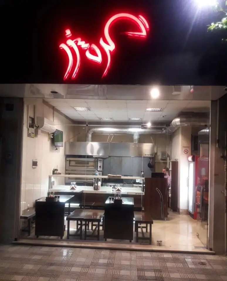 نمای شبانه رستوران فست فود گردسوز با تابلوی نئونی روشن، فضای داخلی مدرن، آشپزخانه اوپن و میز و صندلی‌های چوبی برای مشتریان.