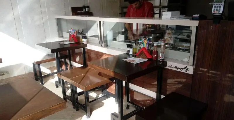 فضای داخلی رستوران گردسوز با میز و صندلی‌های چوبی، پیشخوان آماده‌سازی غذا و آشپز در حال کار. محیطی دنج و مدرن برای مشتریان.