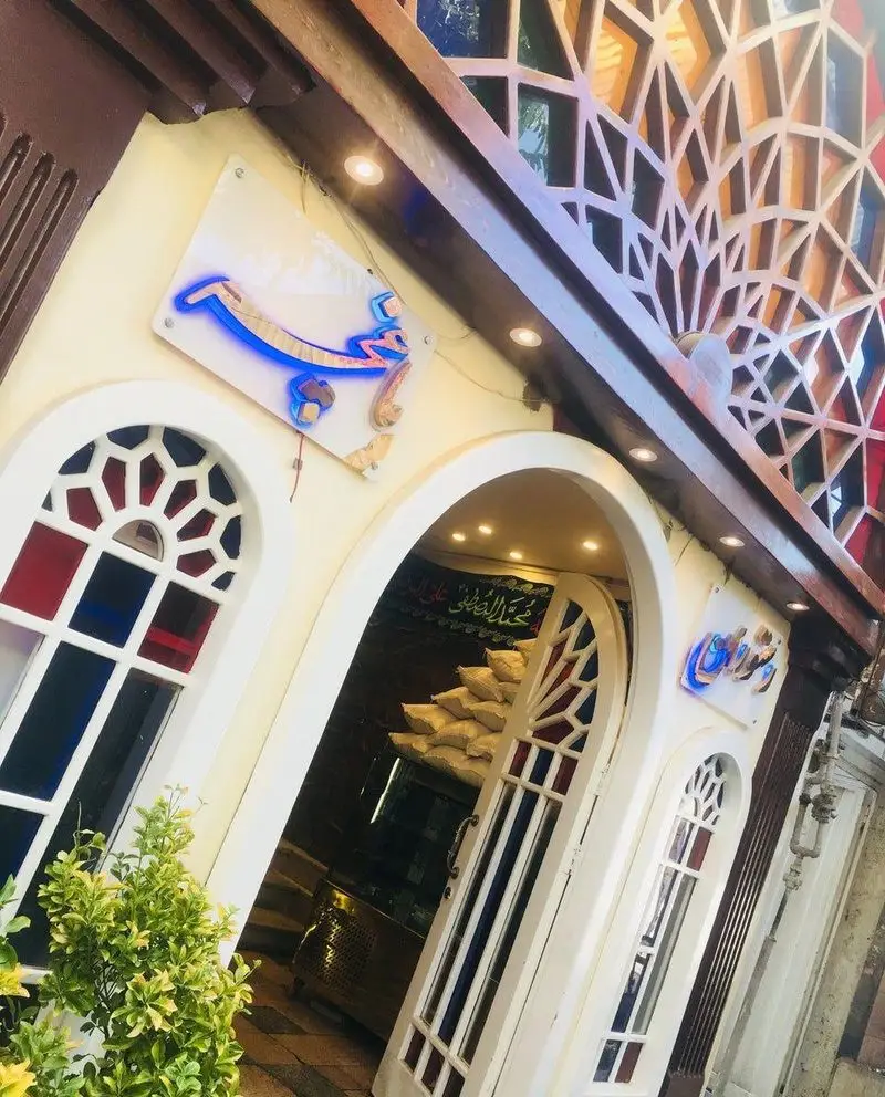 ورودی جذاب رستوران باغچه با تابلوی نئونی آبی، پنجره‌های رنگی و طراحی سنتی، مکانی دلپذیر برای تجربه غذایی لذت‌بخش.