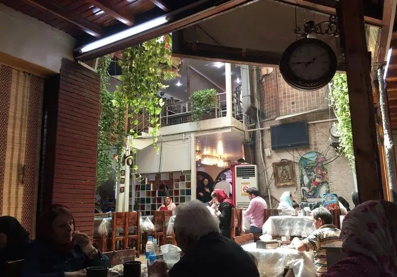 نمایی از رستوران باغچه سنتی و دلنشین با مشتریانی که در حال غذا خوردن هستند، دکوراسیون زیبا و فضای سبز گیاهان آویزان.