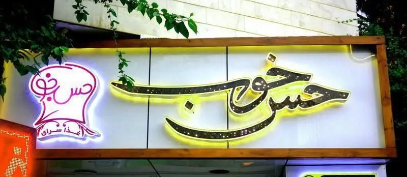 تابلوی سردر تهیه غذا حس خوب (Hess-e Khoub) با حروف درخشان و نماد کلاه سرآشپز، معرف یک کسب و کار موفق در زمینه خوراک.