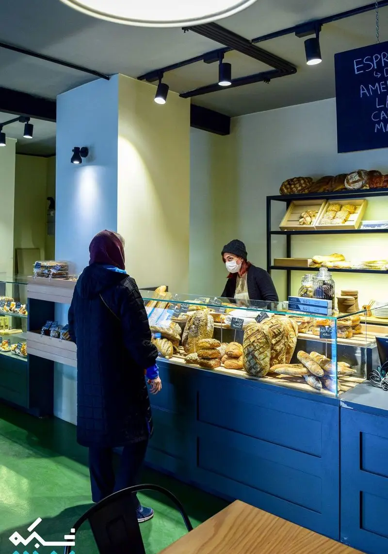 کافه نانوایی دوبار: مشتری در حال انتخاب نان‌های تازه و متنوع از ویترین. نمایشگاهی از بهترین محصولات پخت و شیرینی در محیطی دلپذیر.