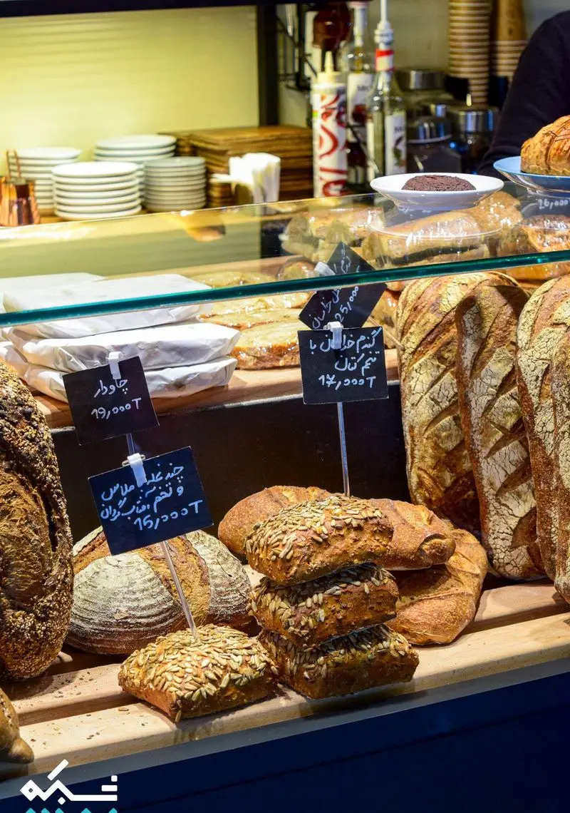 نمایش باشکوه نان‌های تازه دست‌ساز و محصولات متنوع کافه نانوایی دوبار؛ دکون پر از نان چاودار، چند غله و گندم خالص با تخم کتان در کافه‌ای مدرن.