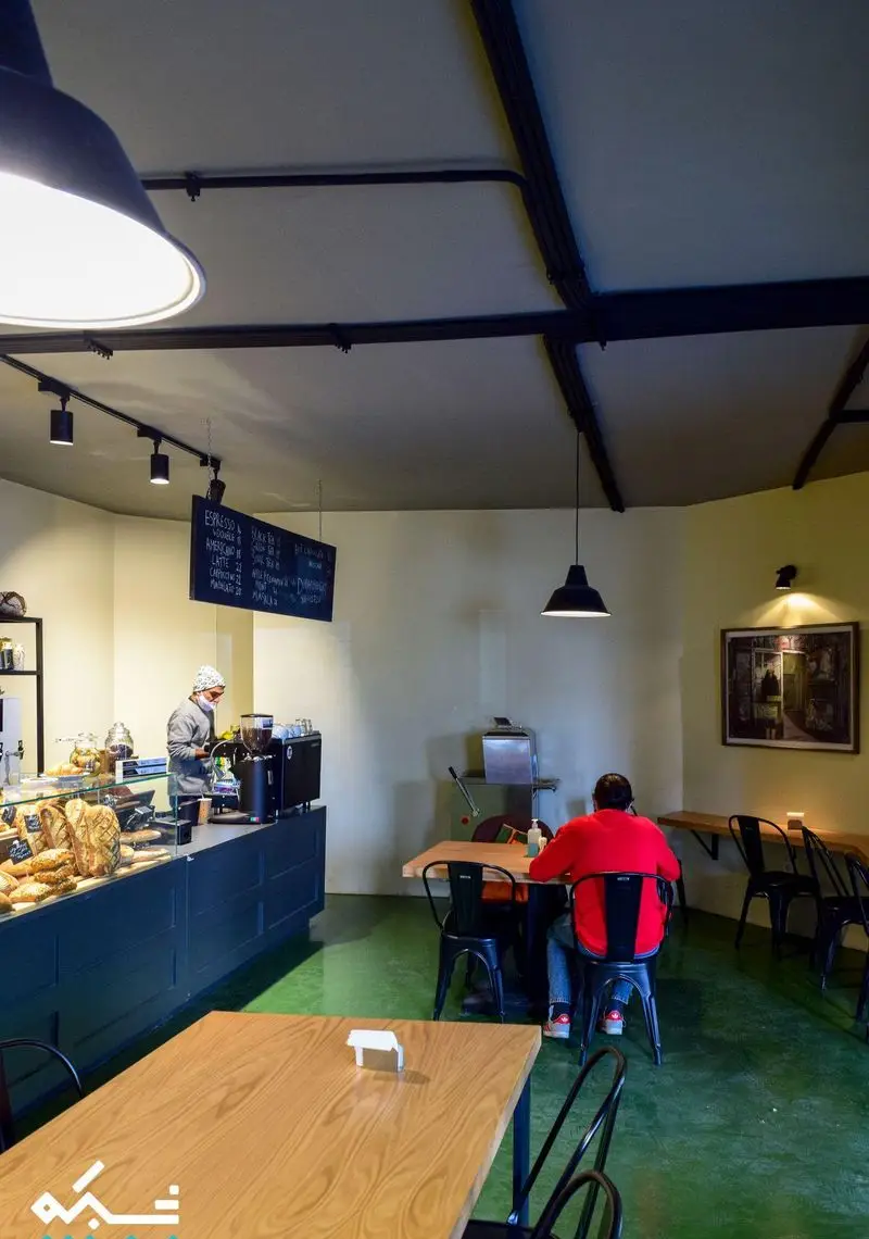 نمای داخلی کافه نانوایی دوبار، با پیشخوان پر از نان تازه، باریستایی در حال آماده‌سازی قهوه و مشتریان در محیطی آرام و مدرن.