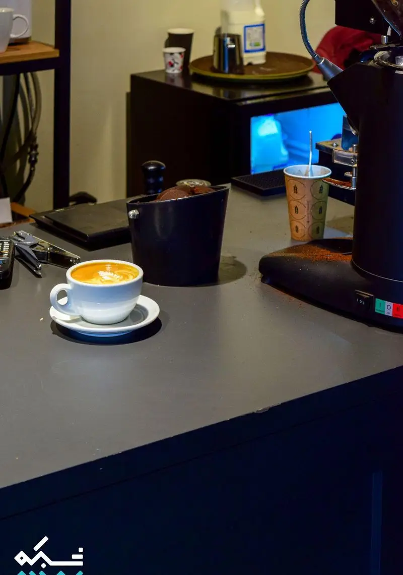 تصویری از میز کار باریستای کافه نانوایی دوبار با فنجان قهوه لاته آرت، دستگاه اسپرسو و تجهیزات آماده‌سازی نوشیدنی.