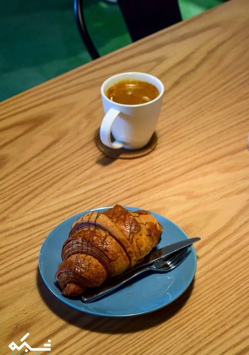 قهوه اسپرسو در فنجان سفید و کروسان شکلاتی لذیذ با کارد و چنگال روی بشقاب آبی در کافه نانوایی دوبار، برای شروع یک روز عالی.