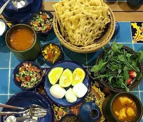 میز رنگارنگ غذای سنتی ایرانی در رستوران پنج دری شامل نان، آش، سبزی تازه، سالاد شیرازی، لیمو و دو نوع خورش با ظروف آبی