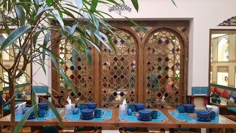 نمایی زیبا از فضای داخلی رستوران و کافه سنتی پنج دری با معماری اصیل ایرانی، ارسی‌های چوبی، شیشه‌های رنگی و میزهای چیده شده با ظروف آبی.