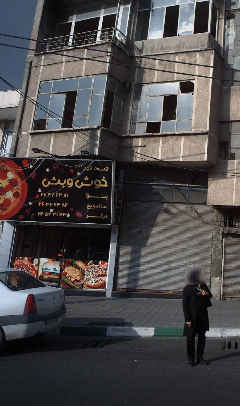 تصویری از فست فود خوش و بش در یک خیابان شهری، با تابلو حاوی پیتزا، ساندویچ و برگر و ویترین غذای آماده.