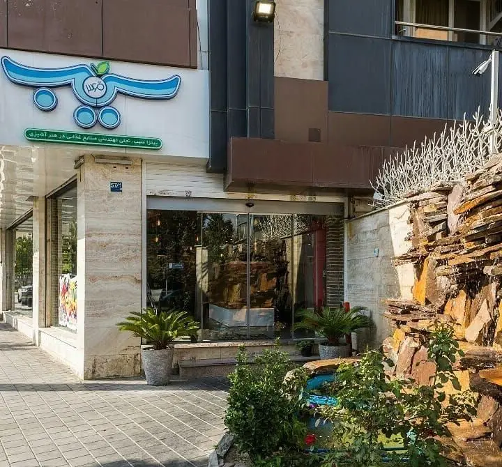 نمای بیرونی جذاب شعبه پیتزا سیب ۳۶۰ با تابلوی آبی رنگ، ورودی شیشه‌ای، آبشار سنگی و فضای سبز. مکانی عالی برای پیتزا و کباب.