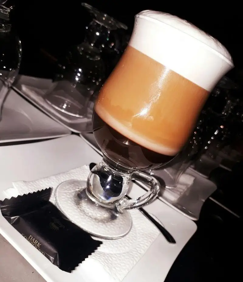 یک فنجان قهوه لایه‌ای فوم‌دار با سه رنگ مختلف و شکلات تلخ در کنار آن، آماده سرو در کافه رستوران برج آزادی، مناسب برای استراحت.