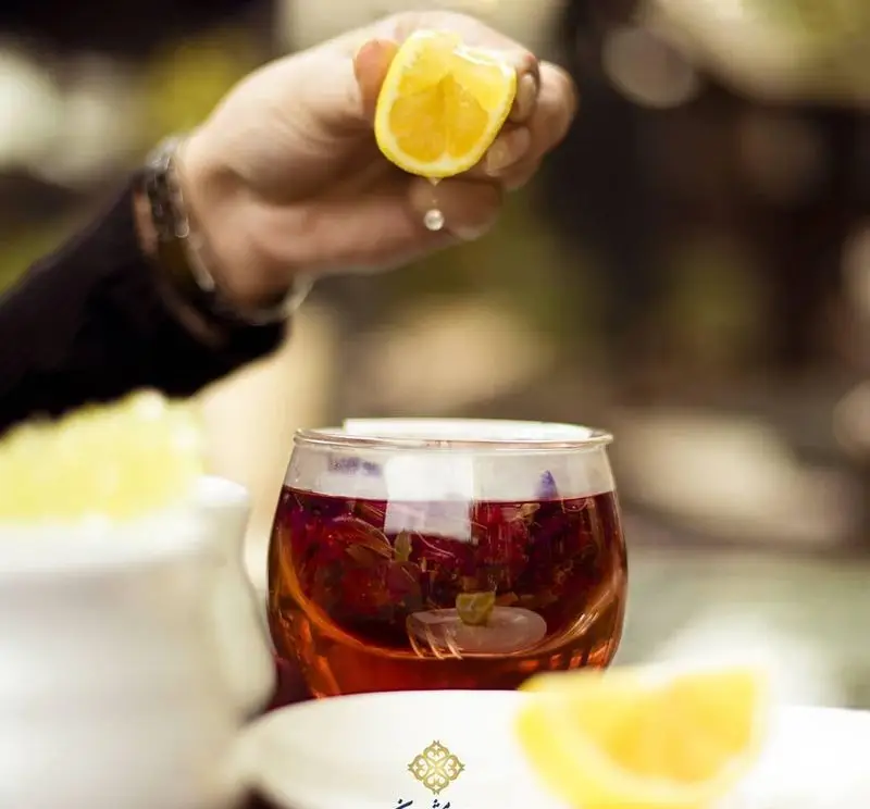فشردن لیموی تازه با دست در لیوان دمنوش اصیل رستوران ایرانی شاندیز ماهان رویال. تجربه‌ای عالی از نوشیدنی‌های سنتی.