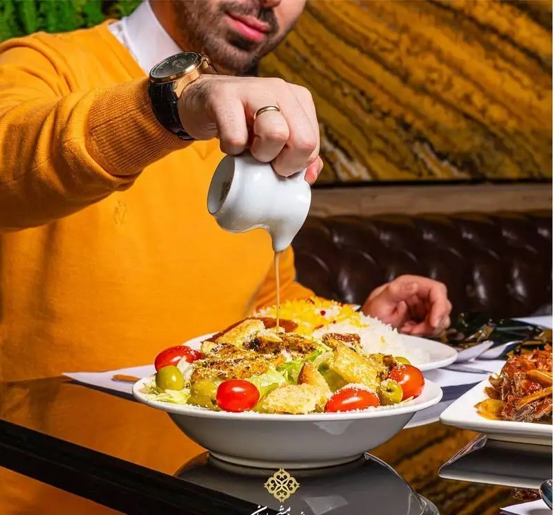 مشتری در رستوران ایرانی شاندیز ماهان رویال، در حال سرو سس روی سالاد خوشمزه، همراه با غذاهای اصیل ایرانی برای تجربه غذایی دلنشین.