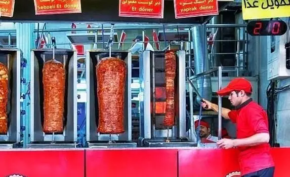 یک آشپز ماهر در حال برش زدن گوشت دنر کباب ترکی تازه و داغ از سیخ‌های چرخان. صحنه‌ای از یک رستوران فست فود دنر آماده سرویس دهی.