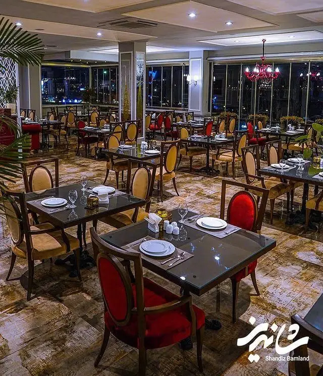 فضای داخلی لوکس و دنج رستوران شاندیز بام‌لند در شب با میزهای چیدمان شده و منظره زیبای شهر، آماده پذیرایی از میهمانان.