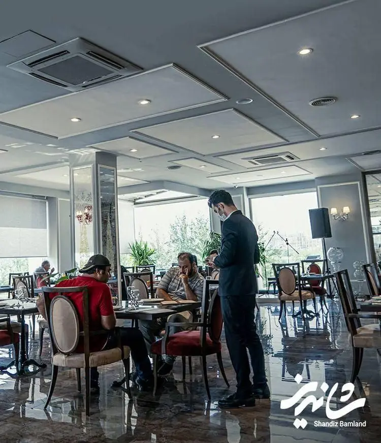 فضای دلنشین رستوران شاندیز بام‌لند با دکوراسیون شیک، گارسون در حال سرویس‌دهی به مشتریان و آماده سازی برای تجربه غذایی لذت‌بخش.