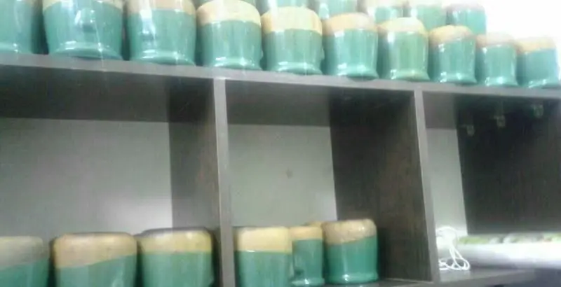 قفسه های منظم پر از ظروف سرامیکی دو رنگ (سبز و کرم) در آشپزخانه رستوران ایرانی و دیزی تندوری، نشانگر کیفیت و نظم.