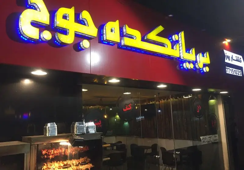 رستوران بریانکده جوجه در شب با تابلوی نئون درخشان. مرغ بریان و کباب‌های تازه در حال پخت روی گریل چرخشی.