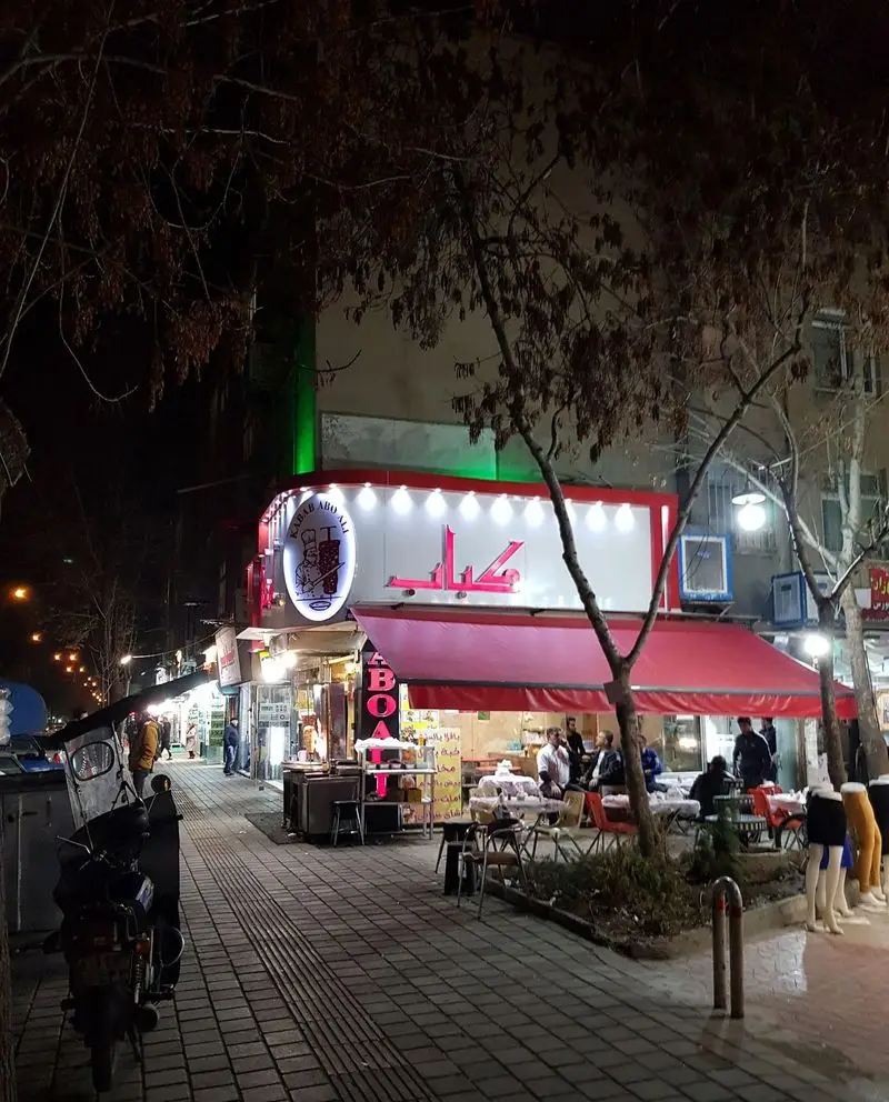 نمای شبانه رستوران کباب ابوعلی، با تابلوی روشن، سقف قرمز و مشتریانی که در فضای باز غذای حلال ایرانی صرف می‌کنند.