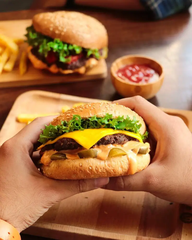 همبرگر شیلا: یک برگر لذیذ با پنیر، کاهو و سس مخصوص در دست، با سیب‌زمینی و برگر دیگر در پس‌زمینه.