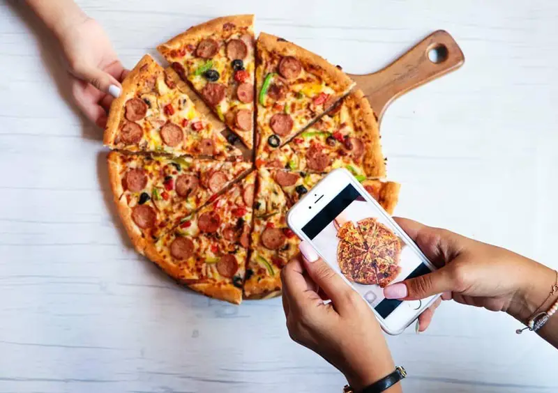 تصویری از پیتزای داغ و تازه فست فود شیلا با تاپینگ سوسیس، فلفل دلمه ای و زیتون که یک نفر اسلایسی برمی‌دارد و دیگری با موبایل عکس می‌گیرد.