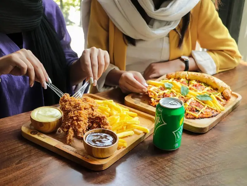 دو نفر در حال صرف غذا در فست فود شیلا: مرغ سوخاری ترد، سیب زمینی سرخ کرده، پیتزای خوشمزه و نوشابه خنک. تجربه‌ای دلپذیر.